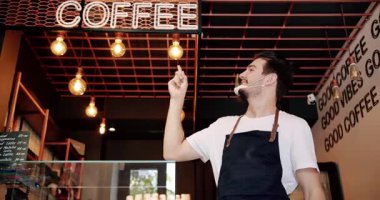 Positive young bearded male barista in white t shirt apron and reusable face mask smiling and looking at camera while standing and point to the inscription coffee in modern coffee house.