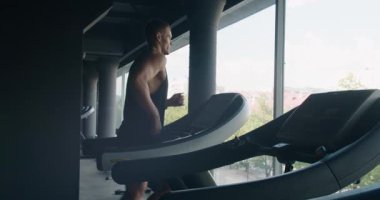Kararlı genç bir adam koşu bandında koşar, şehir manzaralı iyi donanımlı bir spor salonunda spor yapmaya odaklanır. Modern Spor Salonunda Koşu bandına Odaklanmış Genç Adam