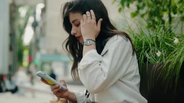 Açık havada otururken akıllı telefonunu kullanan gündelik kıyafetli genç bir kadın. Cihazına odaklanmış, kentsel bir ortamda sakinlik ve rahatlama hissi yayıyor. Akıllı Telefon Kullanan Genç Kadın