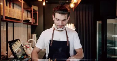 Beyaz tişörtlü, önlüklü, sakallı, modern kafe tezgahında aperatif servisi yaparken gülümseyen ve kameraya bakan genç bir barista.
