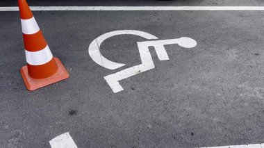 Tekerlekli sandalye sembolü ile işaretlenmiş engelliler için ayrılmış park yeri. Turuncu bir trafik konisi bölgeyi kapatıyor. Engelli sürücüler için erişim, kapsayıcılık ve park kısıtlamaları.