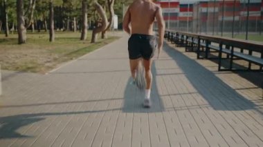 Güneşli bir parkta kaldırımlı bir yolda koşan üstsüz bir adam yeşil ağaçların arasında zindelik ve canlılık ve açık hava stadyumunu gösteriyor. Güneşli Şehir Parkı 'nda Patikada Koşan Üstsüz Adam' ın Arka Görünümü