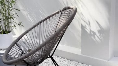 Gri bir Acapulco sandalyesinin rahatlatıcı görüntüsü modern ve minimalist beyaz bir avluda, dekoratif beyaz taşlar ve bir saksı bambu bitkisi....