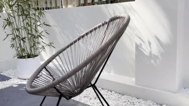 Zarif bir Acapulco sandalyesi ve beyaz tencerede bambu bitkisi olan rahatlatıcı bir açık hava sahnesi. Bahçe alanı beyaz duvarları ve pürüzsüz beton zeminiyle modern. Yaşam tarzı ve tasarım temaları için mükemmel.