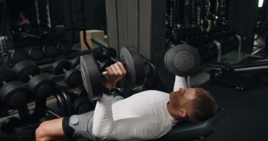 Atletik teçhizatlı odaklanmış genç bir adam, iyi donanımlı bir spor salonunda ağırlık kaldırırken ağır dambılları kaldırır. Modern Spor Salonunda Aptal Benç Baskısı Yapan Genç Adam