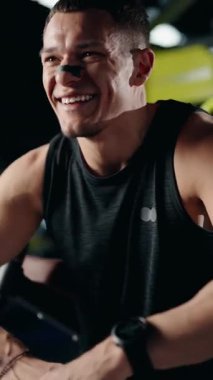 Formda, mutlu bir adam spor salonundaki antrenmanı sırasında gülümser. Fitness izleyicisini kontrol etmek için ara veriyor. Spor kıyafetleri ve burun bandı takıyor. fitness ve sağlık.
