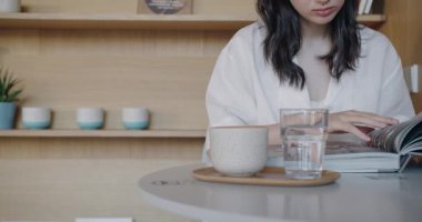 Genç bir kadın şık bir kafede, yanında kahve fincanı ve bir bardak su olan parlak bir dergi okuyor. Sakin ve modern ortam onun dikkatini ve boş zamanını arttırıyor..