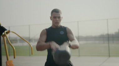 Motive olmuş genç bir erkek fitness meraklısı açık hava ortamında bir çaydanlık çanı kaldırır, güç ve kararlılık yayar. Kettlebell ile Açık Hava Egzersizine Odaklanmış Genç Adam