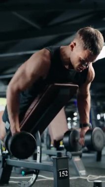 Kas yığını bir adam modern bir spor salonunda dambıl kürek çekiyor. Ağırlıkları odaklanmış kararlılıkla kaldırıyor, güç ve zindelik gösteriyor. Fiziksel kontrollü hareketlerinde sağlıklı bir yaşam tarzı olduğu belli..