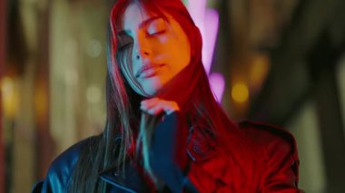 Lüks kadın portresi. Neon ışıkları. Arka plan bulanık. Sokaktaki doğal güzellik kraliçesi. Ağır çekim. Parlak deri ceketli çekici bir kadın neon ışıklarıyla gece caddesinde duruyor..