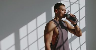 Parlak bir stüdyoda antrenman yaparken atletik bir adam susuzluğunu gideriyor, gölge düşürüyor. Stüdyo Çalışması sırasında Su İçen Genç Atletik Adam