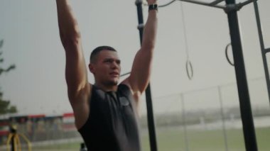 Odaklanmış genç bir adam, açık hava fitness parkında jimnastik halkaları kullanarak yoğun bir çalışma yapıyor. Açık havada jimnastik yapan kendini adamış adam.