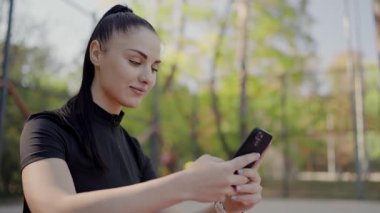 Odaklanmış genç bir kadın spor seansından sonra güneşli bir parkta akıllı telefonunu kontrol ediyor. Sportif kıyafetler giyiyor ve rahatlamış, egzersiz sonrası tavırları var. Sunlit Park 'ta Akıllı Telefon Kullanan Genç Kadın