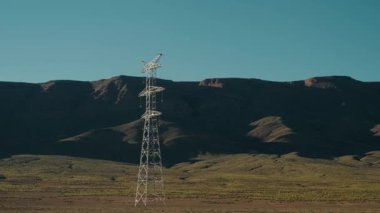 Bir elektrik direği Fas çölünde geniş, dağlık bir zemin üzerinde dimdik duruyor. Engebeli arazi ve açık mavi gökyüzü doğanın kesişmesini sembolize eden güçlü bir kontrast oluşturuyor.