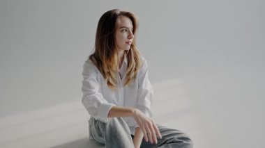 Beyaz gömlek giyen, zarif bir gülümsemesi olan genç bir kadının portresi, parlak bir stüdyo ortamında gündelik ama kendine güvenen bir stil sergiliyor. Beyaz Gömlekli Genç Kadın Stüdyoda Zarif Poz Veriyor