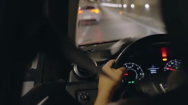 Gece bir tünelden geçerek. Sürücü eli, karanlıkta yol alırken direksiyonda görünür. Gösterge paneli ışıkları iç mekanı aydınlatıyor, hız gösteriyor.