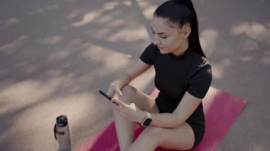 Spor kıyafetli genç bir kadın açık havada pembe yoga minderinde oturmuş, akıllı telefonunu kontrol ediyor. Yanında bir su şişesi var. Dışarıdaki Spor Salonu 'nda Telefonu Kontrol Eden Genç Kadın