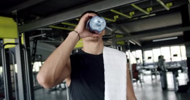 Siyah kolsuz tişörtlü odaklanmış genç bir sporcu, iyi donanımlı, geniş bir spor salonunda yaptığı çalışmadan sonra suyla kendini tazeler. Modern Spor Salonunda Su İçen Fit Genç Adam