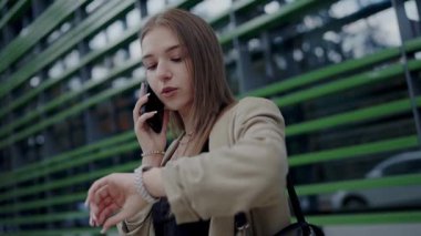 Bir telefon görüşmesi yaparken saatini kontrol eden odaklanmış genç bir kadının portresi, modern mimariyle kentsel bir ortamda duruyor. Şehirde Telefonda Konuşan Genç Kadın Zamanı Kontrol Ediyor