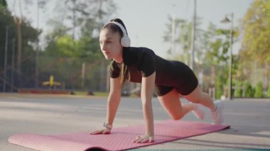 Kulaklıklı, dinamik yoga pozisyonunda, açık havada pembe bir minder üzerinde duran odaklanmış genç bir kadın spor ve dengeyi gösteriyor. Dışarıda Dinamik Yoga Pozu Veren Genç Kadın