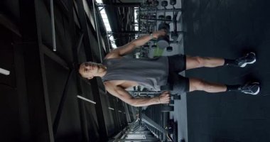 Modern Jimnastik Ortamı 'nda odaklanmış adam eğitim pazıları. Genç atletik bir adam modern bir spor salonunda dambılları kaldırıyor, yoğun bir şekilde egzersiz rutinine odaklanıyor. Dikey video.