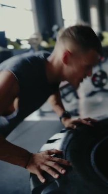 Kas yığını bir adam spor salonunda yoğun bir antrenman yapıyor. Güç antrenmanının bir parçası olarak büyük bir lastiği itiyor. Odaklanmış ve kararlı. Kaslı adam çalışıyor. Dikey