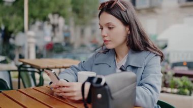 Odaklanmış genç bir kadın cadde kenarındaki bir kafede kahve içerken telefonunu kontrol ediyor ve modern şehir hayatının bir anını yakalıyor. Şehirdeki Outdoor Cafe 'de akıllı telefon kullanan genç bir kadın.