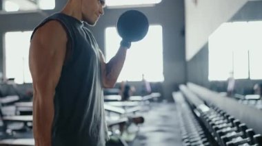 Modern bir spor salonunda dambılları kaldıran odaklanmış bir adam, güç antrenmanı ve formda olduğunu gösteriyor. Spor salonu boş ağırlık ile donatılmıştır ve iyi ışıklandırılmış, sağlıklı bir yaşam tarzı sağlar..