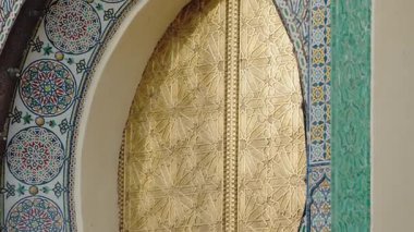 Renkli bir mozaik kemer içinde ayarlanmış karmaşık geometrik desenlerle süslü altın bir kapı. Fas mimarisinin zanaatkarlığı ve sanatsallığı bu detaylı yakın planda sergileniyor..