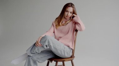 Pembe Süveterli Genç Kadın Tahta Sandalyede Gelişigüzel Poz Veriyor. İnce bir yüzü olan genç bir kadın, rahat pembe bir kazak ve şık gri bir pantolon giymiş, tahta bir sandalyeye rahatça oturur..