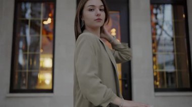 Şık bir trençkot giymiş genç bir kadın, arkasında bir binanın sıcak ışıkları ile bir şehir caddesinde poz vererek başını çeviriyor. Şık Trençkot Giyen Genç Kadın Alacakaranlıkta Poz Veriyor