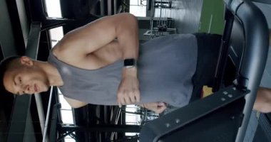 Gri kolsuz tişörtlü kaslı genç bir adam iyi donanımlı bir spor salonunda koşu bandında koşuyor. Kararlılık ve zindelik sergiliyor. Modern Spor Salonu 'nda Koşan Genç Fit Adam. Dikey video.