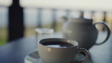 Bir fincan sade kahve ve bir çaydanlık içeren sakin bir görüntü, bir balkonda huzurlu bir sabahı akla getiriyor. Odaklanma yumuşak, sakinliği arttırıyor. Sabah Kahvesi ve Çaydanlık.