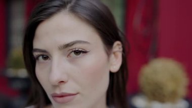 Tarafsız bir yüz ifadesi olan genç bir kadının, bulanık bir geçmişi olan bir şehir ortamında durduğu detaylı bir vesikalık fotoğrafı. Yüz hatlarına ve sakin tavırlarına odaklan..