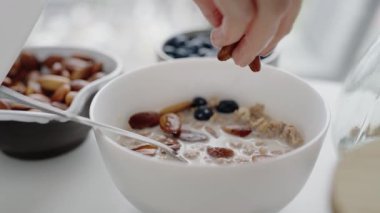 Beyaz bir kase besleyici kahvaltı gevreğine fındık ve böğürtlen ekleyen bir el sağlıklı beslenme alışkanlıklarını ve sağlıklı bir yaşam tarzını teşvik ediyor. Sağlıklı Kahvaltıya Fındık ve Böğürtlen Eklemek