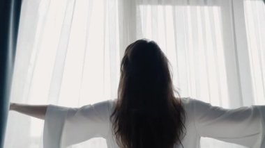 Beyaz cüppeli bir kadın, perdeleri doğal ışık için açıyor. Yeni bir başlangıcı ve yeni bir başlangıcı simgeliyor. Sabah Perdeleri Parlak Doğal Işığa Açan Kadın