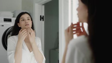 Banyodaki aynaya bakarken beyaz cüppeli bir kadın cilt bakımı yapıyor. Kişisel bakım, güzellik rutini ve sağlıklı deri kavramları. Banyo Aynası Yansıması 'na Cilt Rutini Uygulayan Kadın