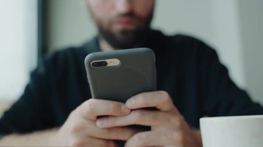 Kafedeyken akıllı telefon kullanan bir adamın yakın çekimi var. Telefonuna bir mesaj yazıyor gibi görünüyor. Sakin ve odaklanmış bir atmosferle kapalı bir ortam. Cafe 'de kahve fincanıyla akıllı telefon kullanan adam.