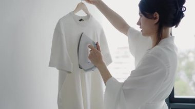 Beyaz cüppeli bir kadın, parlak ve minimalist bir odada beyaz bir gömleği askıda ütülüyor. Minimalist Ayarlarda Kadın Buharı Giysileri Gündüz Penceresinin Yanındaydı