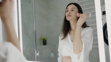 Genç bir kadın modern bir tuvalette günlük cilt bakımı rutininin bir parçası olarak yeşim taşını kullanıyor. Kendine özen ve güzelliğe odaklan. Banyodaki Aynanın Önünde Yüz Pateni Kullanan Kadın