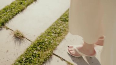 Taş bir yolda yürürken, beyaz bir elbise ve sandalet giymiş, sıcak ve sakin bir atmosferi olan bir kadının yakın çekimi. Bahçede Beyaz Elbise Giyen ve Sandalet Giyen Kadının Yakın Çekimi