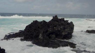 Beyaz okyanus dalgalarının güçlü bir sahnesi, bulutlu bir gökyüzünün altında siyah, engebeli lav kaya oluşumlarına şiddetle çarpıyor. Dramatik Okyanus Dalgaları Rocky Lav oluşumlarına karşı çarpışıyor