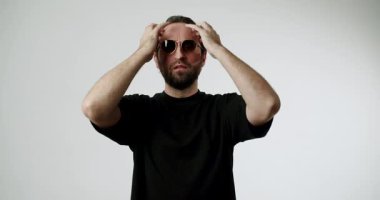 Sakallı ve güneş gözlüklü şık bir adam saçını beyaz bir arka plana göre ayarlar. Kendine güven ve karizma yayıyor, modern stil ve bireysellik hissini yakalıyor..