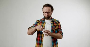 Sakallı ve gözlüklü bir adam beyaz bir plastik ilaç şişesini beyaz bir stüdyo arkaplanına karşı açmaya çalışıyor. Şapkayı bükmeye çalışırken hayal kırıklığına uğramış görünüyor. Bu video zorluk tasvir eder