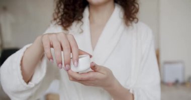 Ellerinde bornoz olan genç bir kadın yüz kremiyle bir şişe açıyor ve evde kokuyor. Beyaz krema kavanozunu açtım. Tedavi gören güzel bayan vücut bakımını nemlendiriyor.,