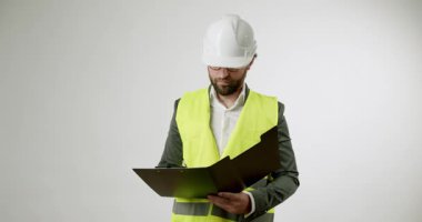 Beyaz şapka ve güvenlik yeleği giymiş bir inşaat denetçisi belgeleri bir panoda gözden geçiriyor. Ciddi ve odaklanmış görünüyor. Basit beyaz bir geçmişin evraklarını kontrol ediyor..