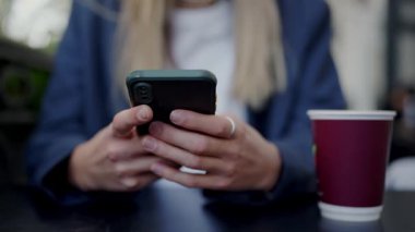 Genç bir kadın şehirdeki bir kafede, önünde kahve fincanı olan akıllı telefonuna odaklanıyor, gelişigüzel bir şehir yaşamını canlandırıyor. Şehir Kafesi 'nde Akıllı Telefon Kullanan Genç Kadın