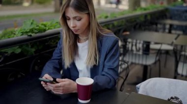 Odaklanmış genç bir kadın, şehir yaşamını ve bağlantısını somutlaştıran bir kahve fincanı olan açık bir kafe masasında akıllı telefonunu kullanıyor. Dışarıdaki Kafe Ayarları 'nda Genç Kadın Mesajlaşıyor