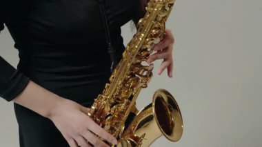 Siyah elbiseli genç bir kadın stüdyoda beyaz arka planda saksafon çalıyor. Saksafon çalan bir bayan müzisyen. Siyah bir gece elbisesi giymiş, sahnede solo yapan bir kadın saksafoncu..