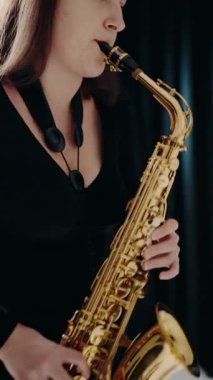 A young woman in black dress plays the saxophone on the black background in studio. Jazz musician playing the saxophone. A female saxophonist wearing a black evening dress performing. vertical